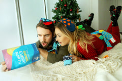 A couple laying on a bed looking with shock into a bag that says the joy of a surprise every month.