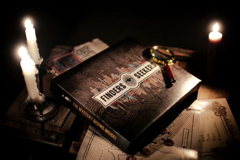 A Finders Seekers Escape Room Game subscription box with a magnifying glass on top and lit candles nearby.