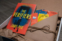 Book titled the Verifiers by Jane Pek resting in box next to book wrapped in brown paper and twine stuffed with items