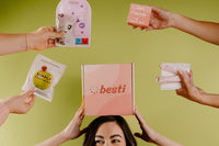 A girl holding a Besti box and surrounded by products that are included in the subscription like snacks, face masks, and organic pads. 