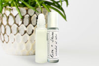 Two bottles of the Rise + Shine Essential Oil Roller standing side by side in front of a potted plant.