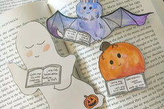 A ghost, pumpkin and vampire bat bookmark all sit on an open hardcover book. Each bookmark is detailed and hand painted.
