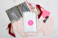 A My Fashion Crate subscription box surrounded by red high heels, a pink blouse, sunglasses and magazines.