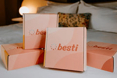 A collection of Besti boxes that are arranged stacked together on a comfy bed with a lamp in the background. 
