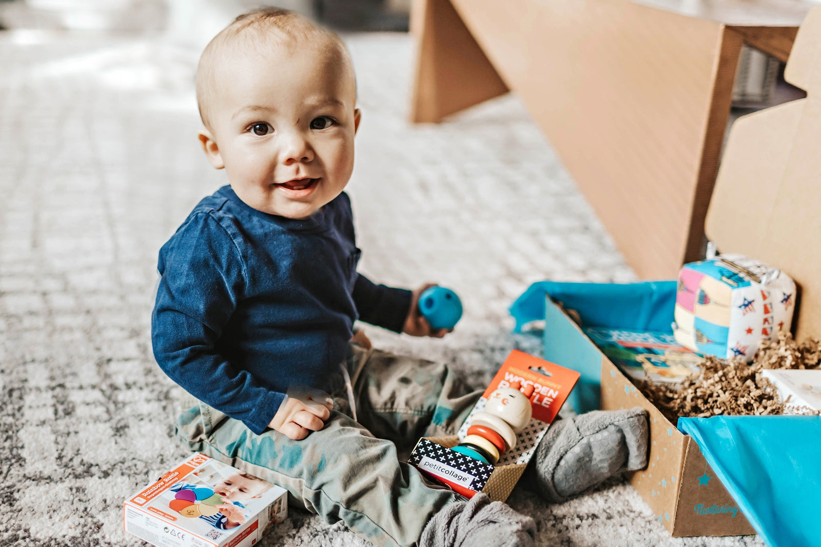 A baby boy sitting in front of a Hoppi subscription box and playing with baby toys from the box.