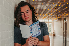 a young adult male learning up against a white brick wall holding a greeting card that says can't stop won't stop and reading the inside