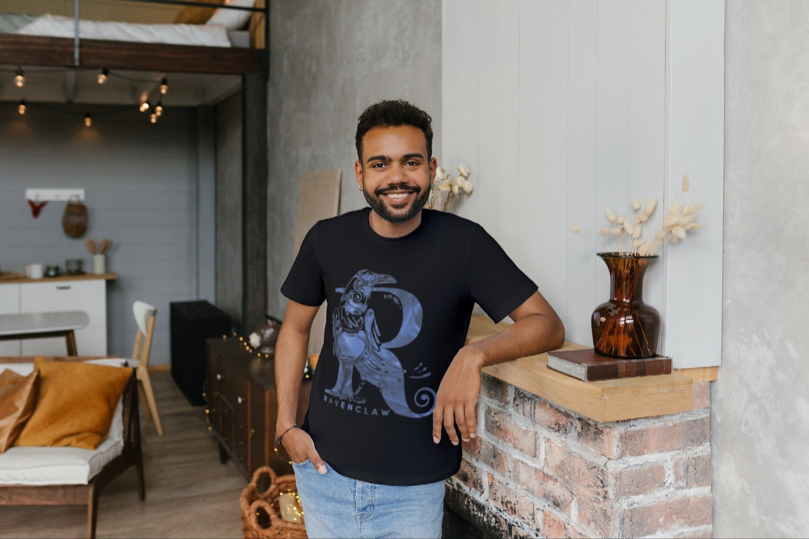 A man leaning against a shelf wearing a black Harry Potter Tshirt with a Raven making the shape of an R.