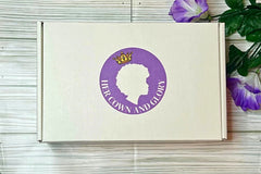 Her Crown and Glory subscription box against wooden-style background placed next to purple flowers  