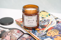 a jarred candle named cartoons and comics sits atop a messy assortment of open comic books. The comics are sitting on a white table.