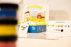 Out of focus paint containers with an open Hello Family kit featuring Printston's Art Studio and additional Color Combo Pad.