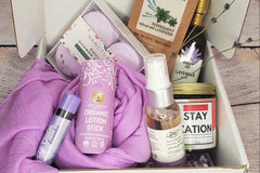 An open subscription box containing a lavender colored scarf, organic lotion stick, shower bombs and lavender body spray.
