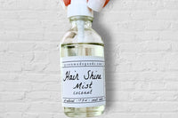 A hand holding the Coconut Hair Shine Mist by Greenmade goods with a white brick wall in the background.