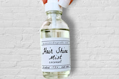 A hand holding the Coconut Hair Shine Mist by Greenmade goods with a white brick wall in the background.