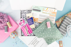Looking down on an open box of IndieStitch. Green and pink fabrics, with thread, fabric ruler grips, and a pin cushion are in the open box.