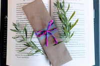 A fantasy romance-themed bookmark wrapped in brown craft paper and adorned with pink and purple ribbons rests on an open book.